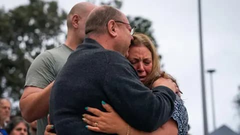 Matilda's father, dressed in a grey pullover, embraces her weeping mother, dressed in a blue and white t shirt