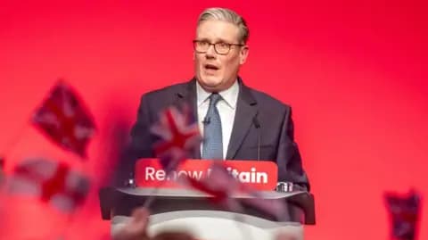 Starmer speaks at a podium in front of a red wall, with people waving Union Jacks in the crowd