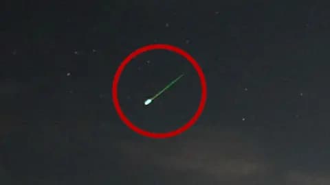 A meteor with a bright green tail streaks through the night sky, with several stars nearby. The meteor is circled with a red overlay.