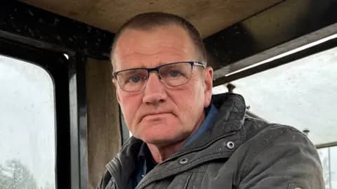 A farmer, a man with short black hair wearing glasses and a coat, sitting in the cab of a tractor on a rainy overcast day.