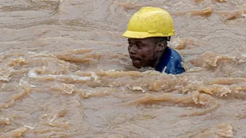 The head of a man in a yellow hardhat is above brown floodwater which is swirling around him. 
