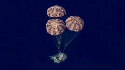 Three red and white parachutes bring the Orion capsule gently into the Pacific Ocean with a splash.