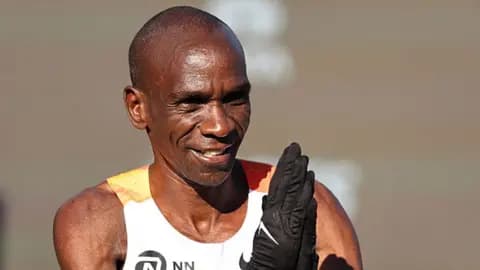 A head and shoulders shot of Eliud Kipchoge in a running vest and holding his gloved hands up in prayer.