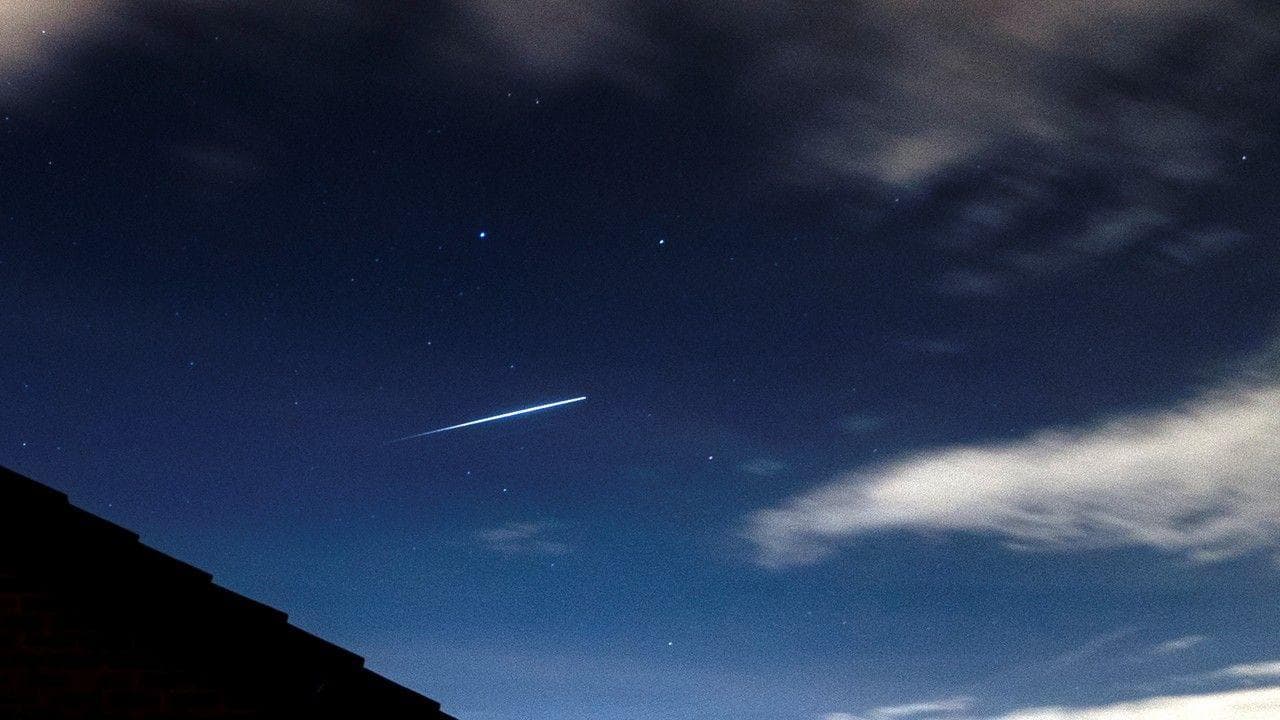 The white streak of a meteor set against the blue night sky with white clouds to the right and above and the outline of a rooftop to the bottom letf