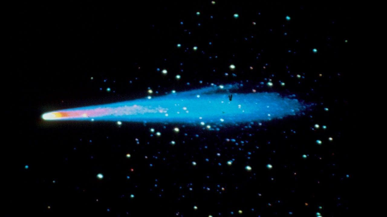 A photo of a comet with a yellow and pink head, and blue tail, streaking across the black background of space punctuated by lights from stars
