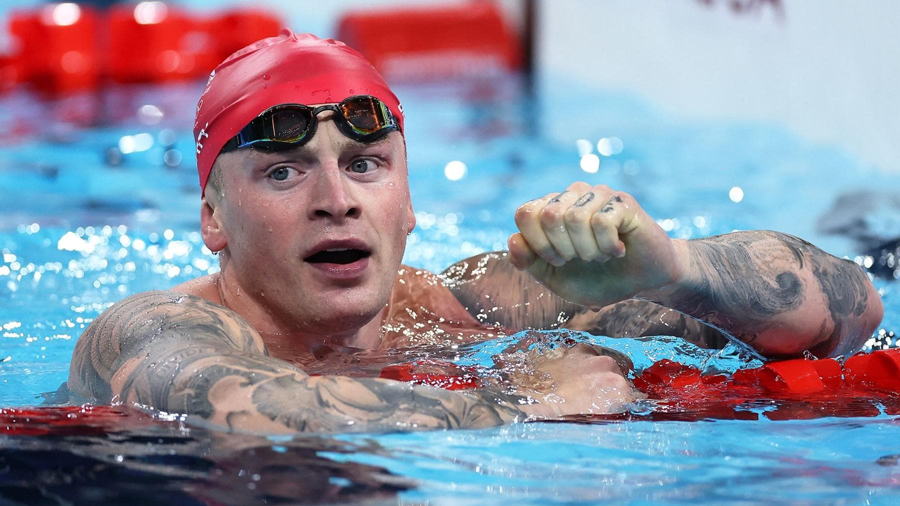 Adam Peaty in the pool wearing a red swimming cap. He is not swimming, he is leaning on a floating lane divider