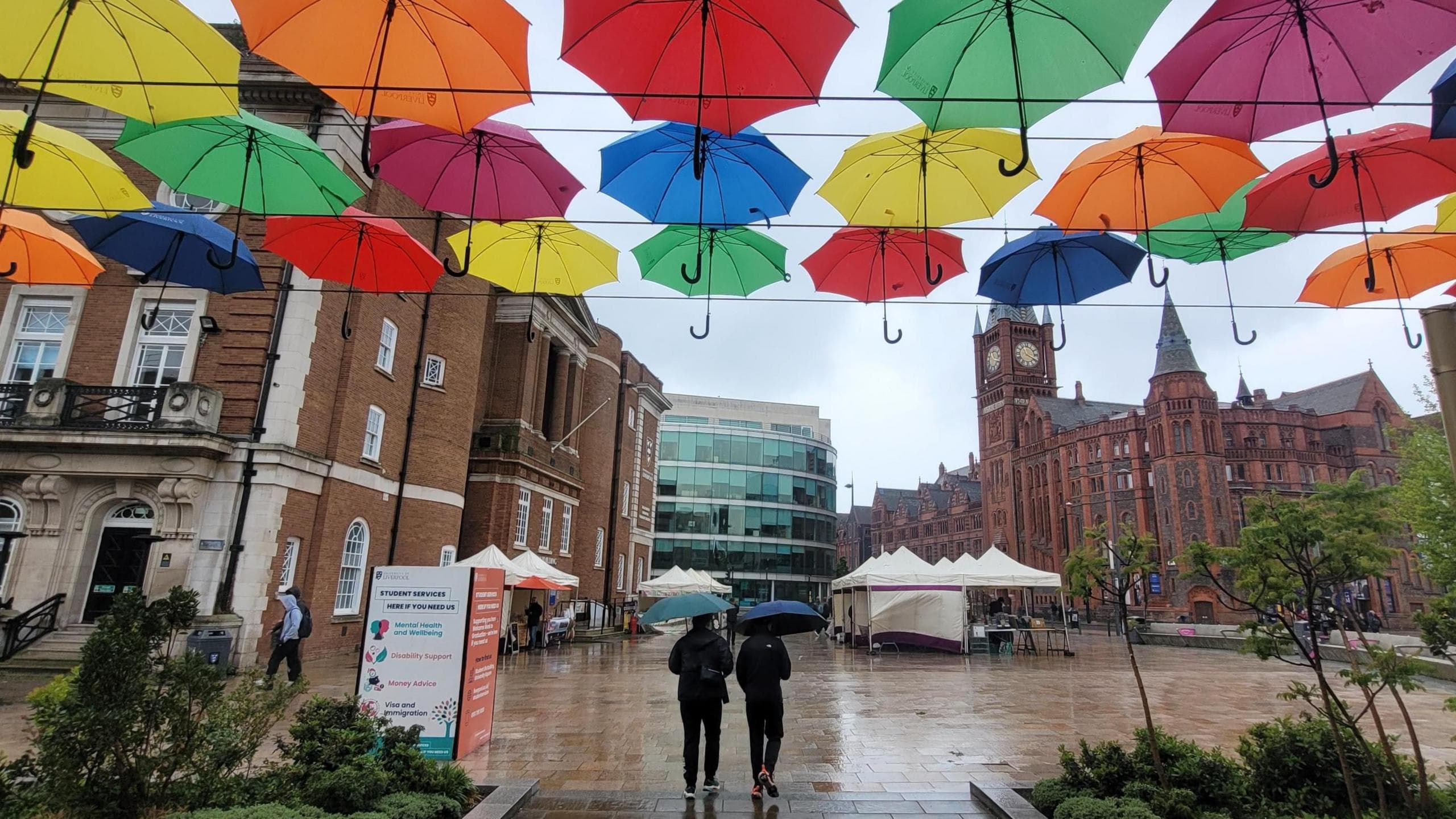 Two people holding umbrellas walk across a rain-soaked city square, with rows of colourful umbrellas suspended overhead