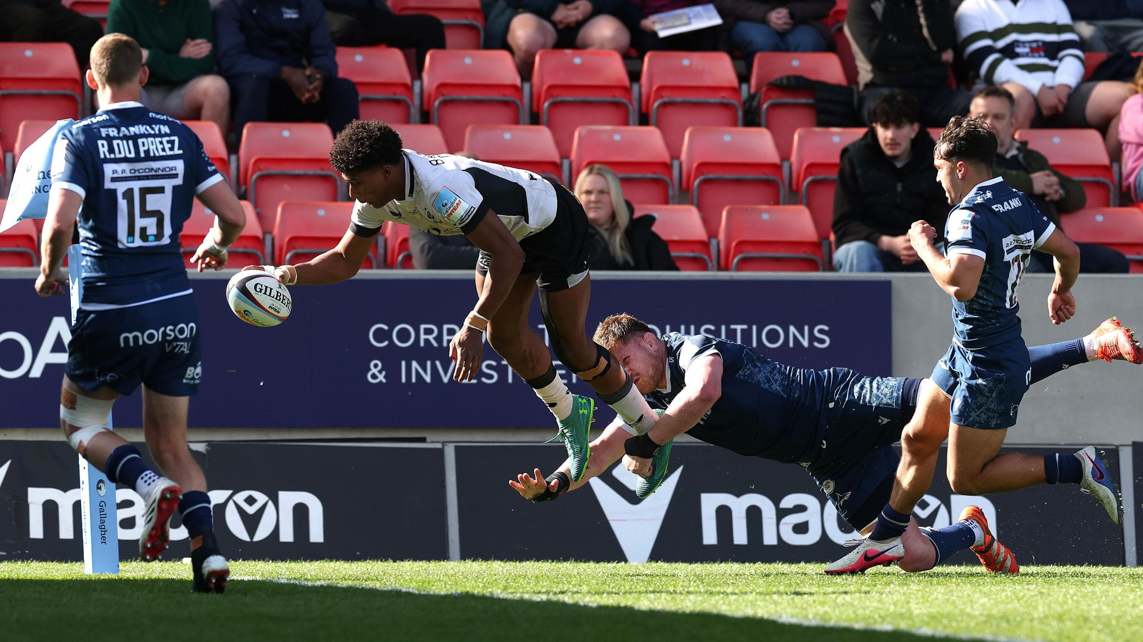 Saracens winger Noah Caluori diving a few feet off the ground to score a try as one Sale player tries to tackle him and two others (one left and one right) watch on powerlessly