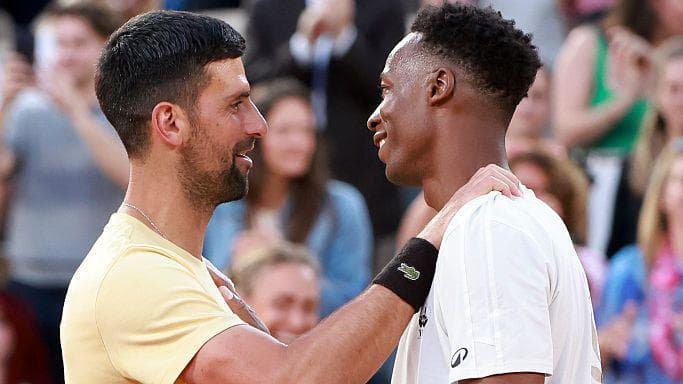 Novak Djokovic, in a pale yellow T-shirt, stands facing Gael Monfils, wearing a white T-shirt, rests his right arm on the Frenchman's shoulder