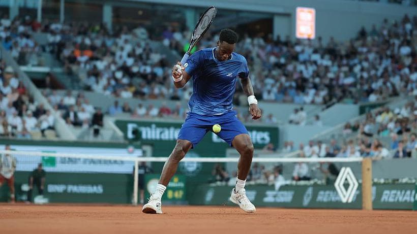 Gael Monfils, wearing a blue T-shirt and shorts, lines up a shot between his legs with the ball between his thighs and his black racquet held in his right hand at head height