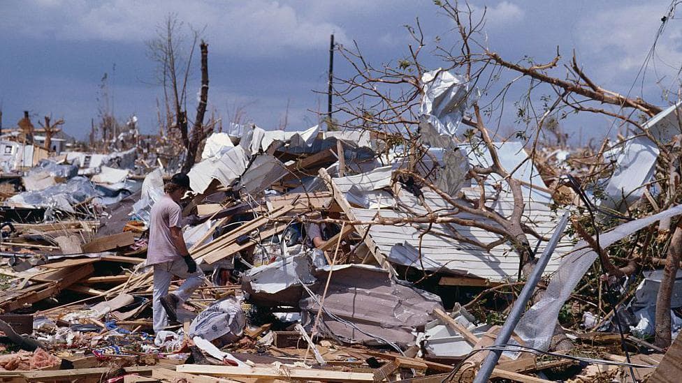 A man surveys the wreckage of buildings and trees with a mix of blue sky and clouds overhead