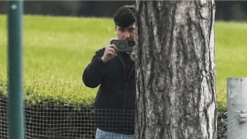 A man in a dark jacket and blue jeans stands partially concealed by a tree trunk and holds a phone up in two hands as if filming and has a white earphone in his right ear