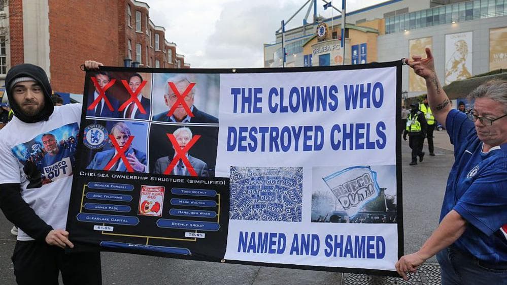 Two Chelsea fans hold up a banner critical of the club's owners, reading "The Clowns Who Destroyed Chels - Named And Shamed"